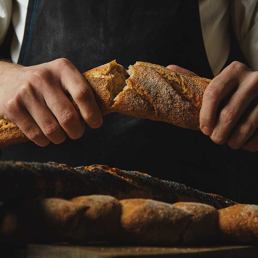 primer-plano-deliciosas-mitades-baguette-organica-manos-hombre-fondo-delantal-negro-mesa-madera-marron